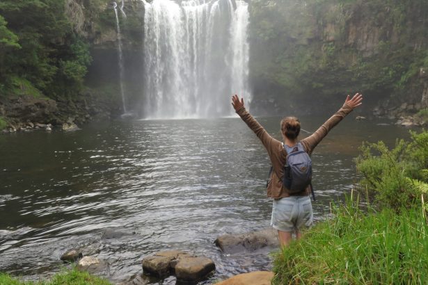 A hiker with her arms raised marveling at Rainbow Falls waterfall. Kerikeri, New Zealand